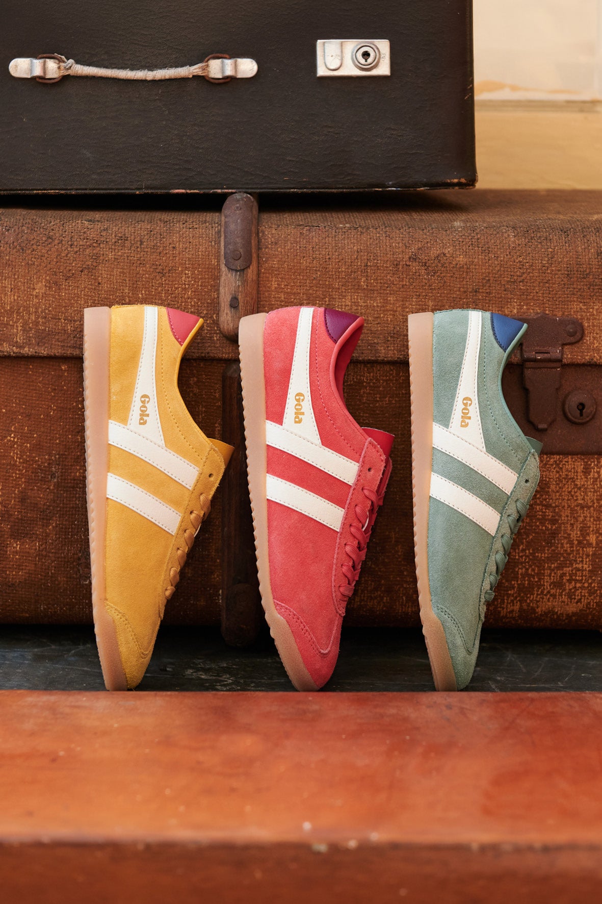 Three Gola trainers in yellow, red, and green, with white stripes, against vintage suitcases