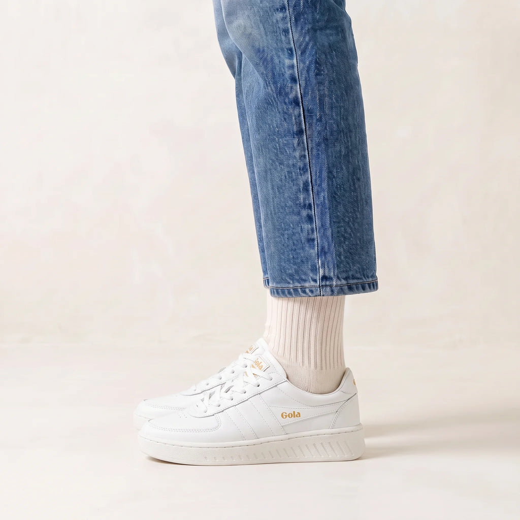 White platform sneakers with gold 'Gola' branding, worn with white socks and blue jeans against a light beige background.