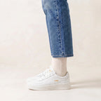 White platform sneakers with gold 'Gola' branding, worn with white socks and blue jeans against a light beige background.