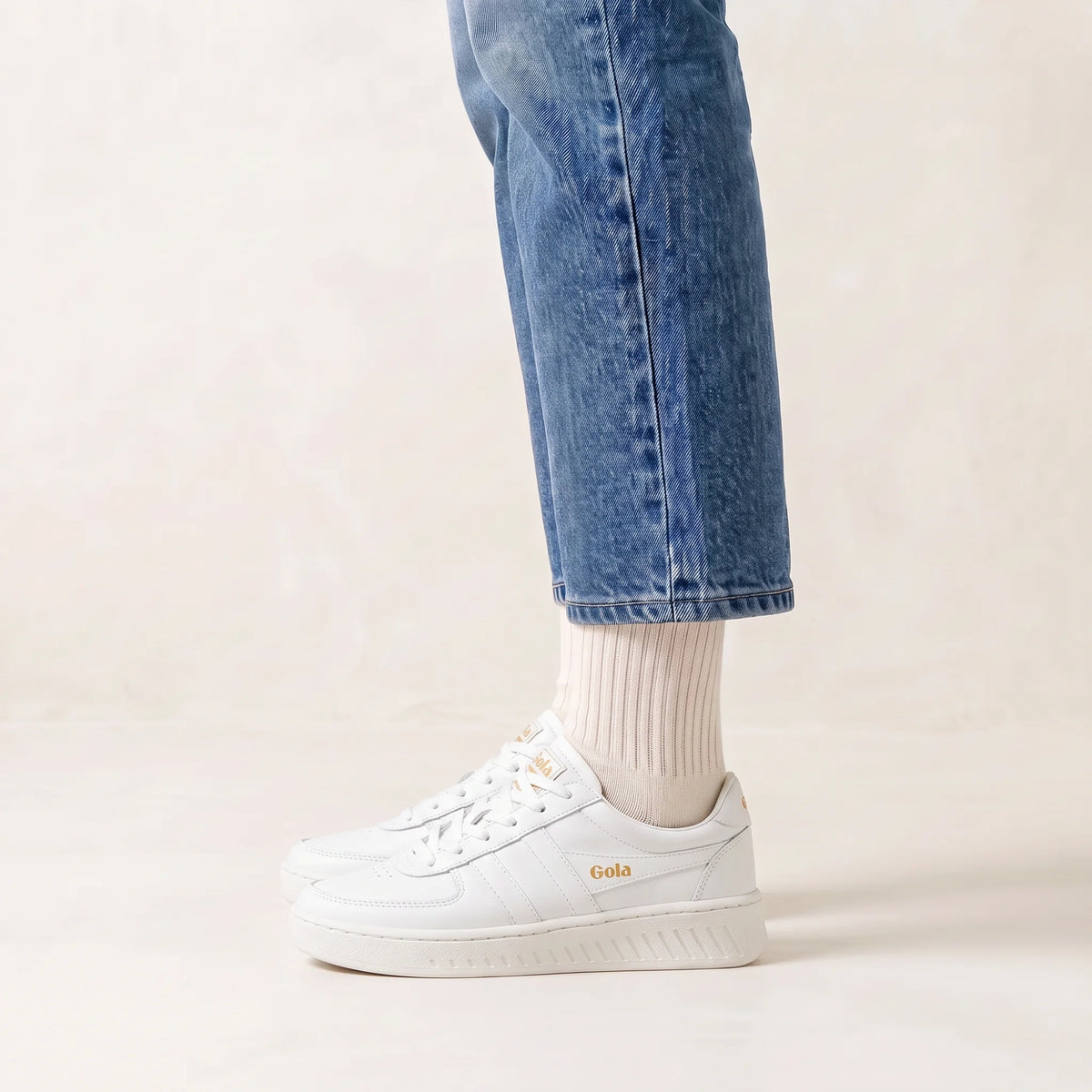 White platform sneakers with gold 'Gola' branding, worn with white socks and blue jeans against a light beige background.