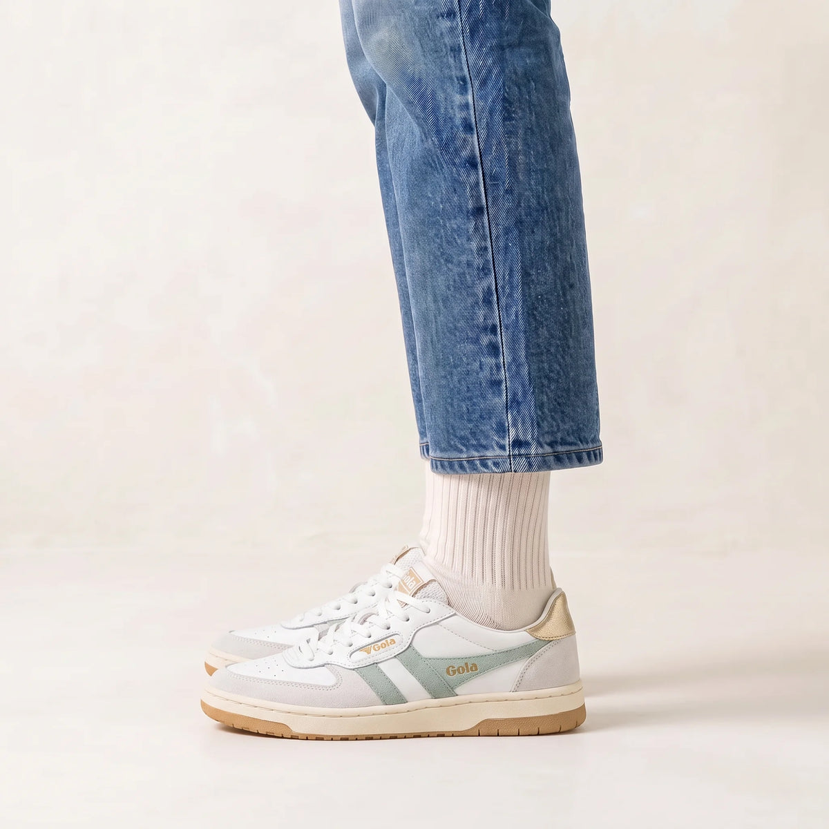 A person wearing light blue jeans, white socks, and white low-top sneakers with sage green side stripes and gold 'Gola' branding, standing against a warm off-white studio background.