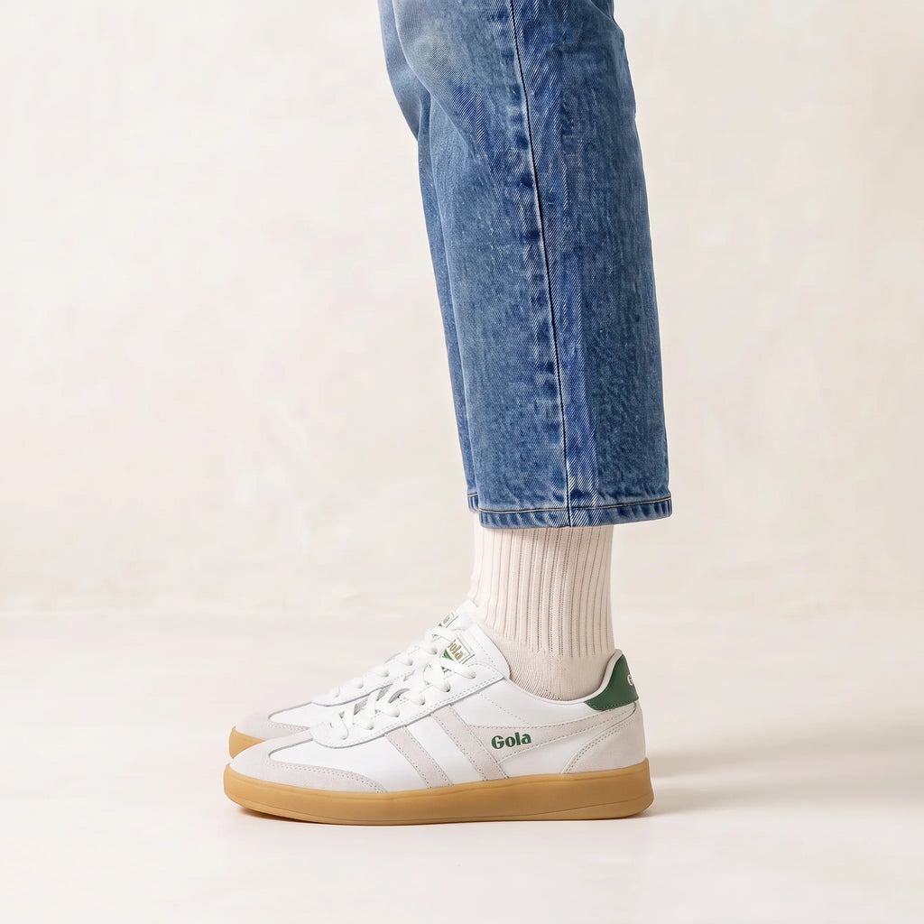 White Gola sneakers with green accents and gum soles, worn with white socks and blue jeans on a light beige background.
