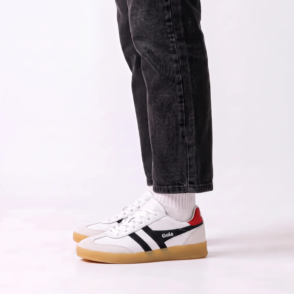 White Gola sneakers with black side stripes, a red heel tab, and a gum sole, shown on a model wearing black jeans and white socks against a white studio background.