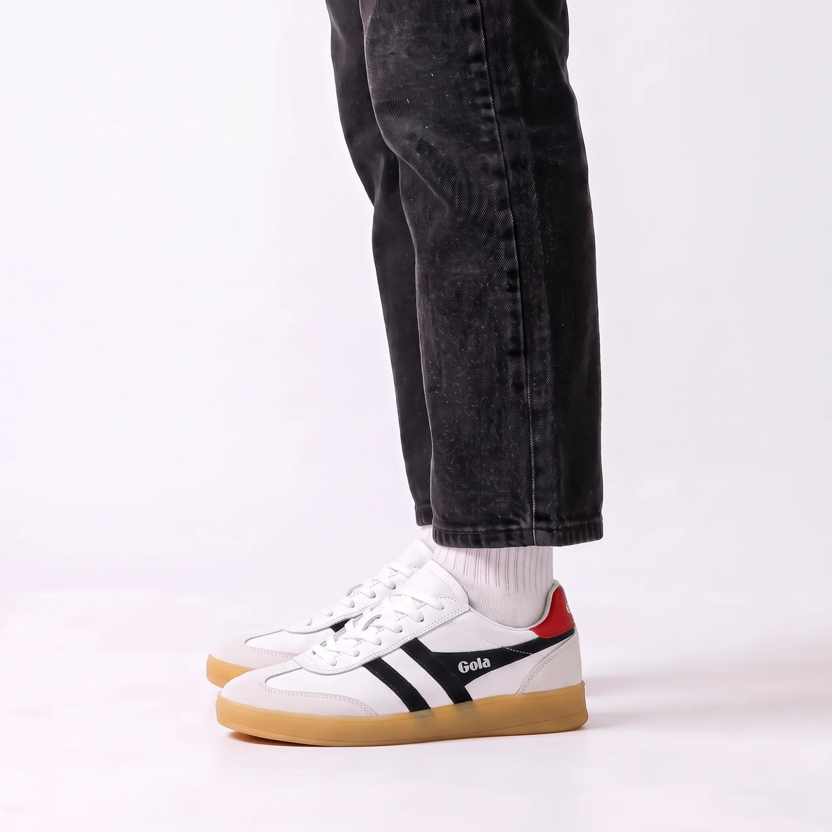 White Gola sneakers with black side stripes, a red heel tab, and a gum sole, shown on a model wearing black jeans and white socks against a white studio background.