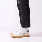 White Gola sneakers with green accents and gum soles, shown on a model wearing black jeans and white socks against a white studio background.