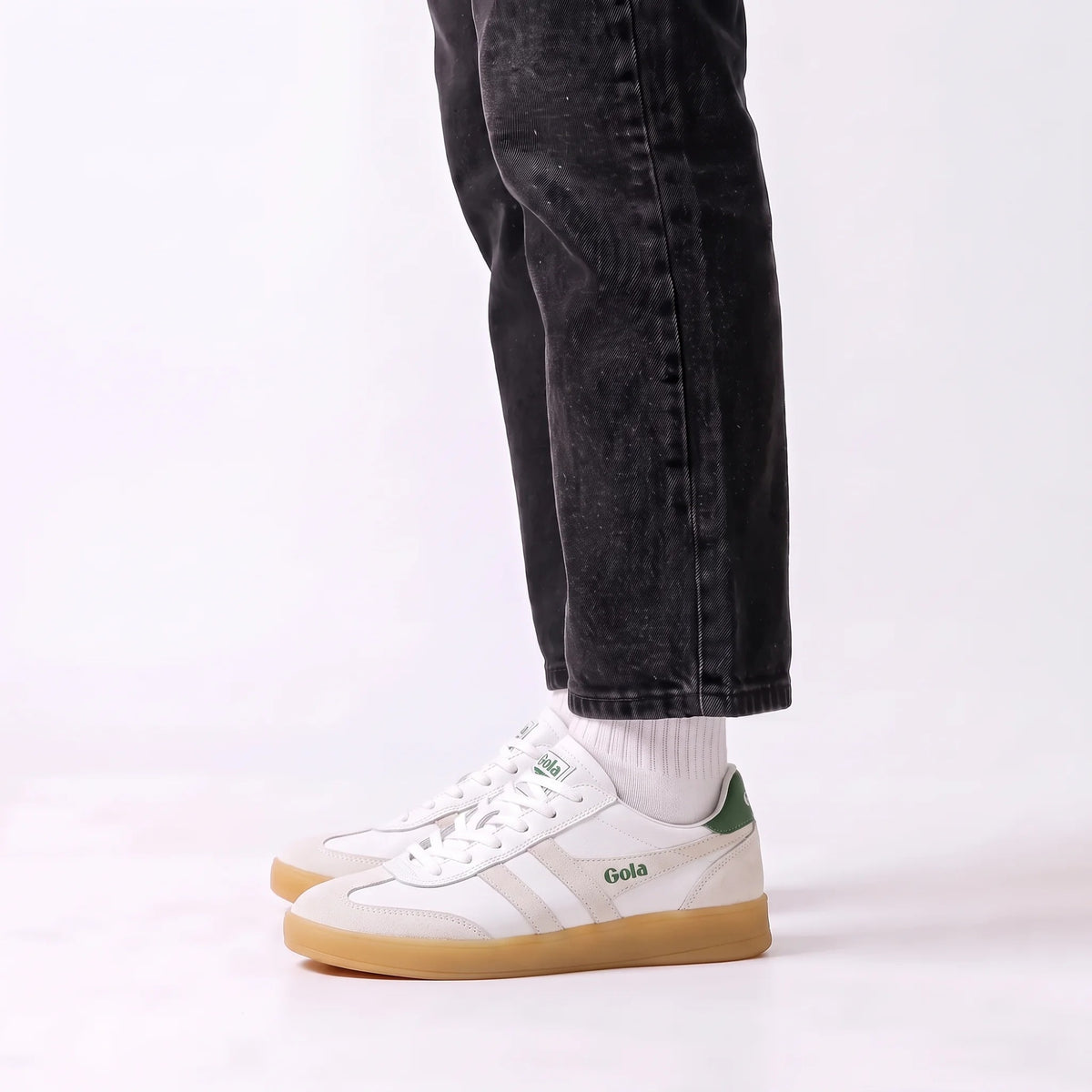 White Gola sneakers with green accents and gum soles, shown on a model wearing black jeans and white socks against a white studio background.