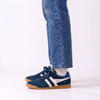 A person wearing blue jeans and white socks, shown from the calves down, wearing a pair of navy blue Gola sneakers with white side stripes and gum soles, against a plain white studio background.