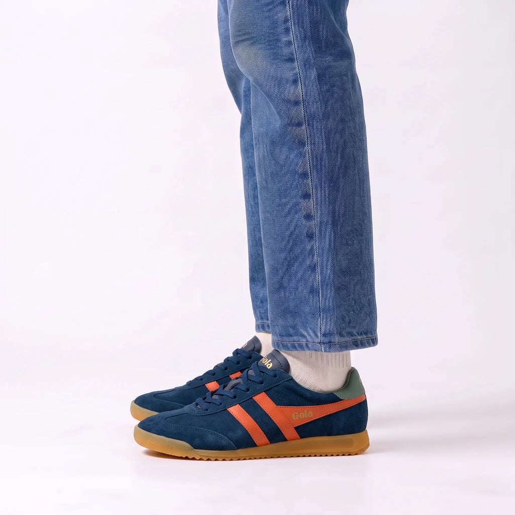 A person wearing blue suede sneakers with orange side stripes and gum soles, paired with blue jeans and white socks, shown from the calves down against a clean white studio background.
