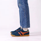 A person wearing blue suede sneakers with orange side stripes and gum soles, paired with blue jeans and white socks, shown from the calves down against a clean white studio background.