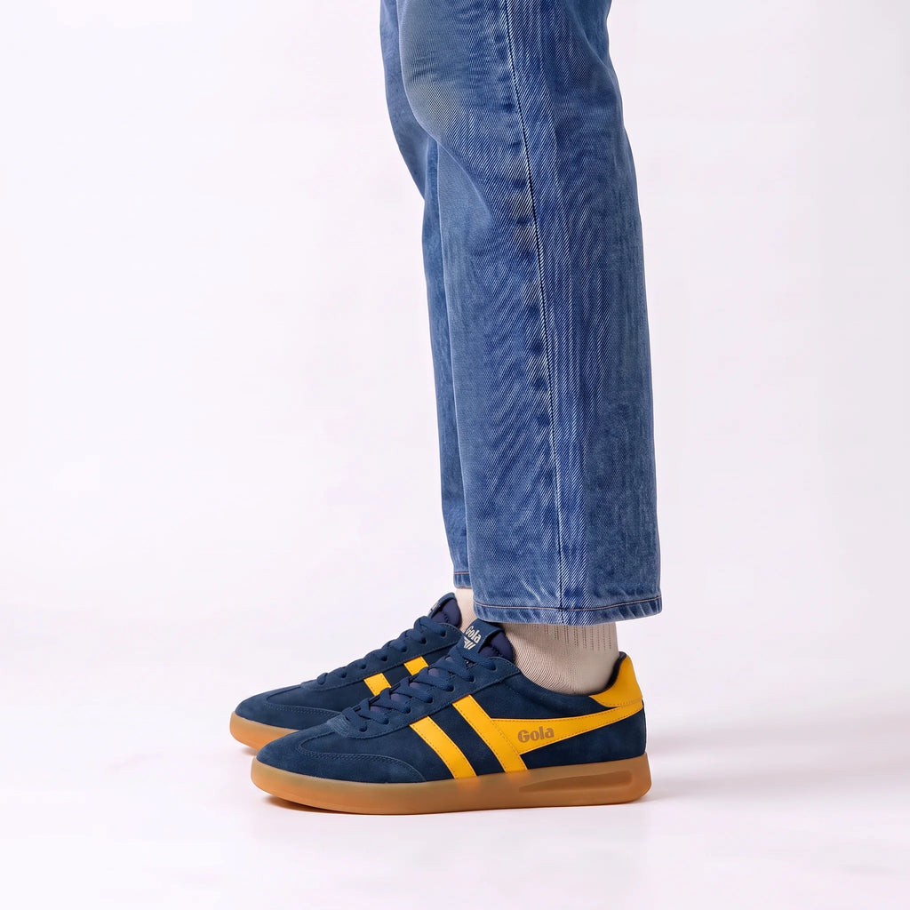 A person wearing blue jeans and navy suede sneakers with yellow stripes and a gum sole, shown from the calves down against a clean white studio background.