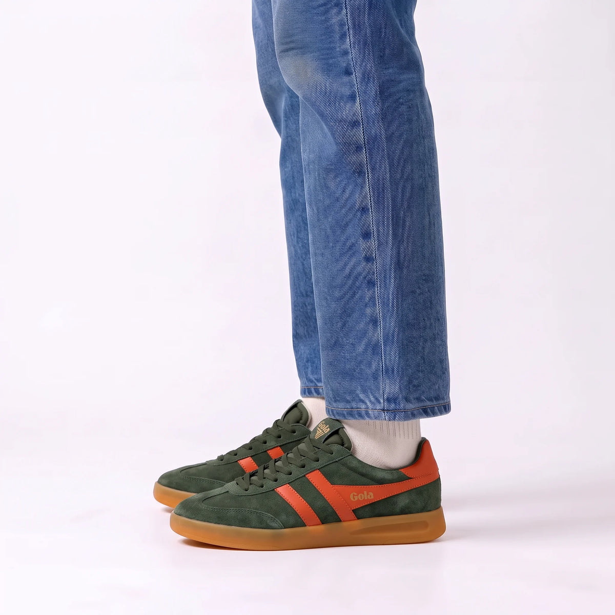 A person wearing green suede Gola sneakers with orange stripes and a gum sole, paired with blue jeans and white socks, shown from the knees down against a plain white studio background.