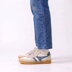 A person wearing blue jeans and off-white Gola sneakers with blue side stripes and a gum sole, shown from the calves down against a clean white studio background.