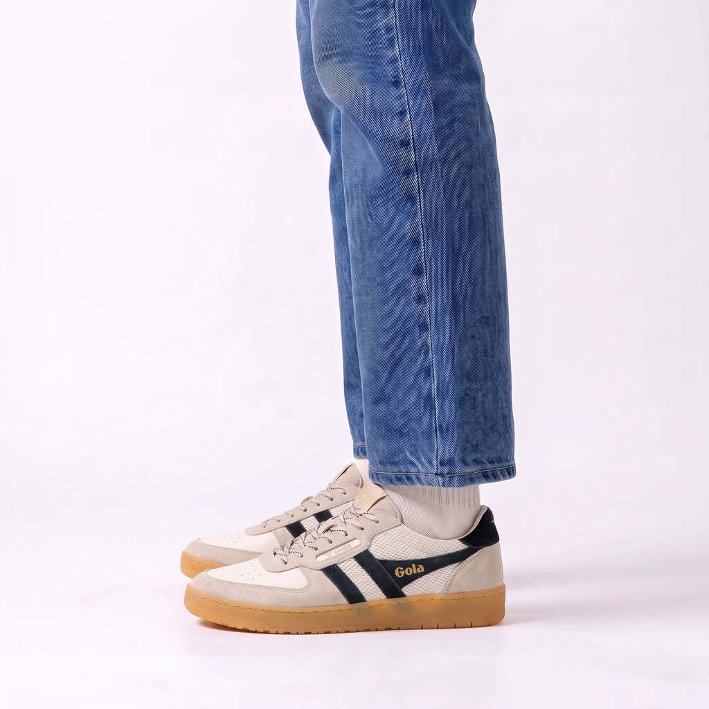A person wearing blue jeans and white socks stands in a studio setting, showing a pair of Gola sneakers. The shoes are primarily beige and white with dark navy stripes and a gum sole.