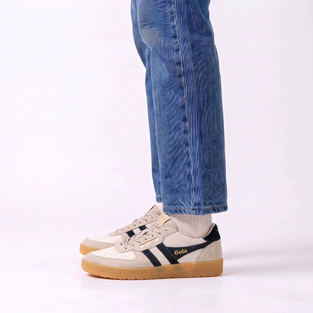 A person wearing blue jeans and white socks stands in a studio setting, showing a pair of Gola sneakers. The shoes are primarily beige and white with dark navy stripes and a gum sole.