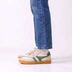 A person wearing blue jeans and white socks with a pair of beige and green Gola sneakers with gum soles, shown from the knees down against a white studio background.