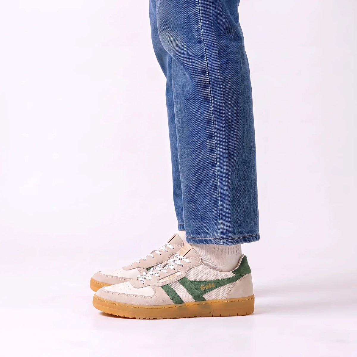A person wearing blue jeans and white socks with a pair of beige and green Gola sneakers with gum soles, shown from the knees down against a white studio background.