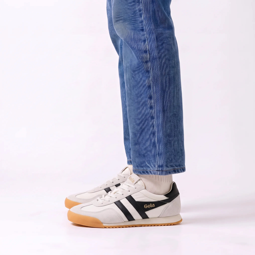 A person wearing blue jeans and white socks with a pair of white Gola sneakers featuring black side stripes and a gum sole, shown from the knees down on a white studio background.