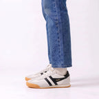 A person wearing blue jeans and white socks with a pair of white Gola sneakers featuring black side stripes and a gum sole, shown from the knees down on a white studio background.