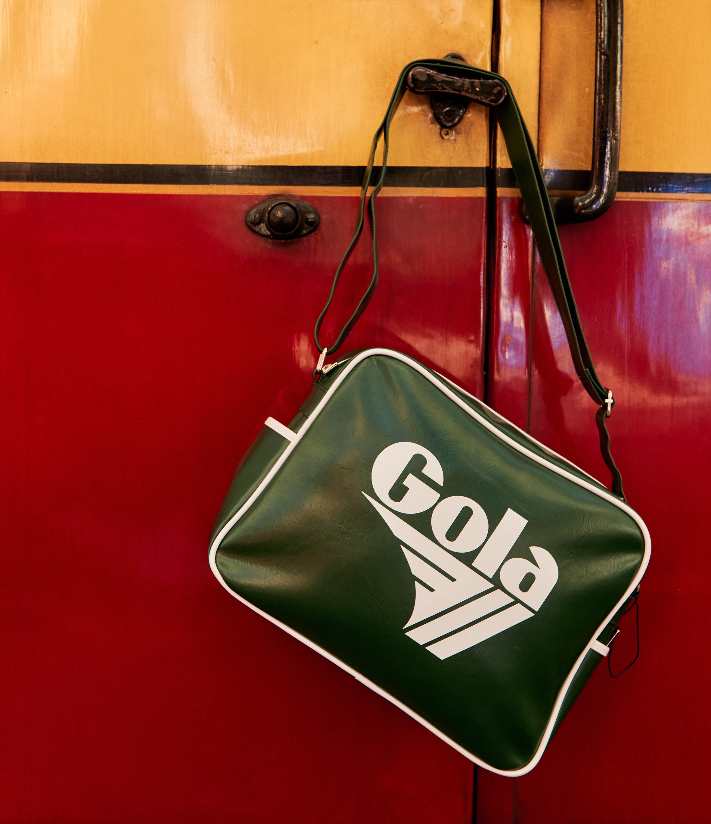 A green vintage-style shoulder bag with white piping and a large white 'Gola' logo, hanging against a red and yellow painted metal background.