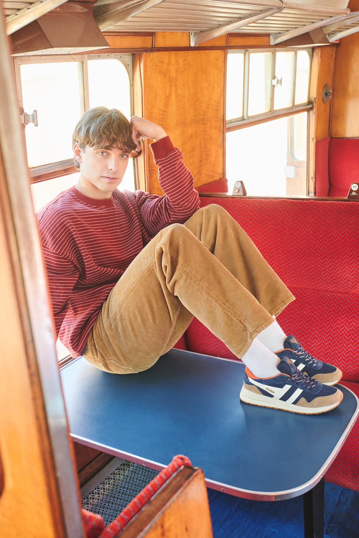 Man in a red striped jumper and tan corduroy trousers sitting on a table in a train carriage