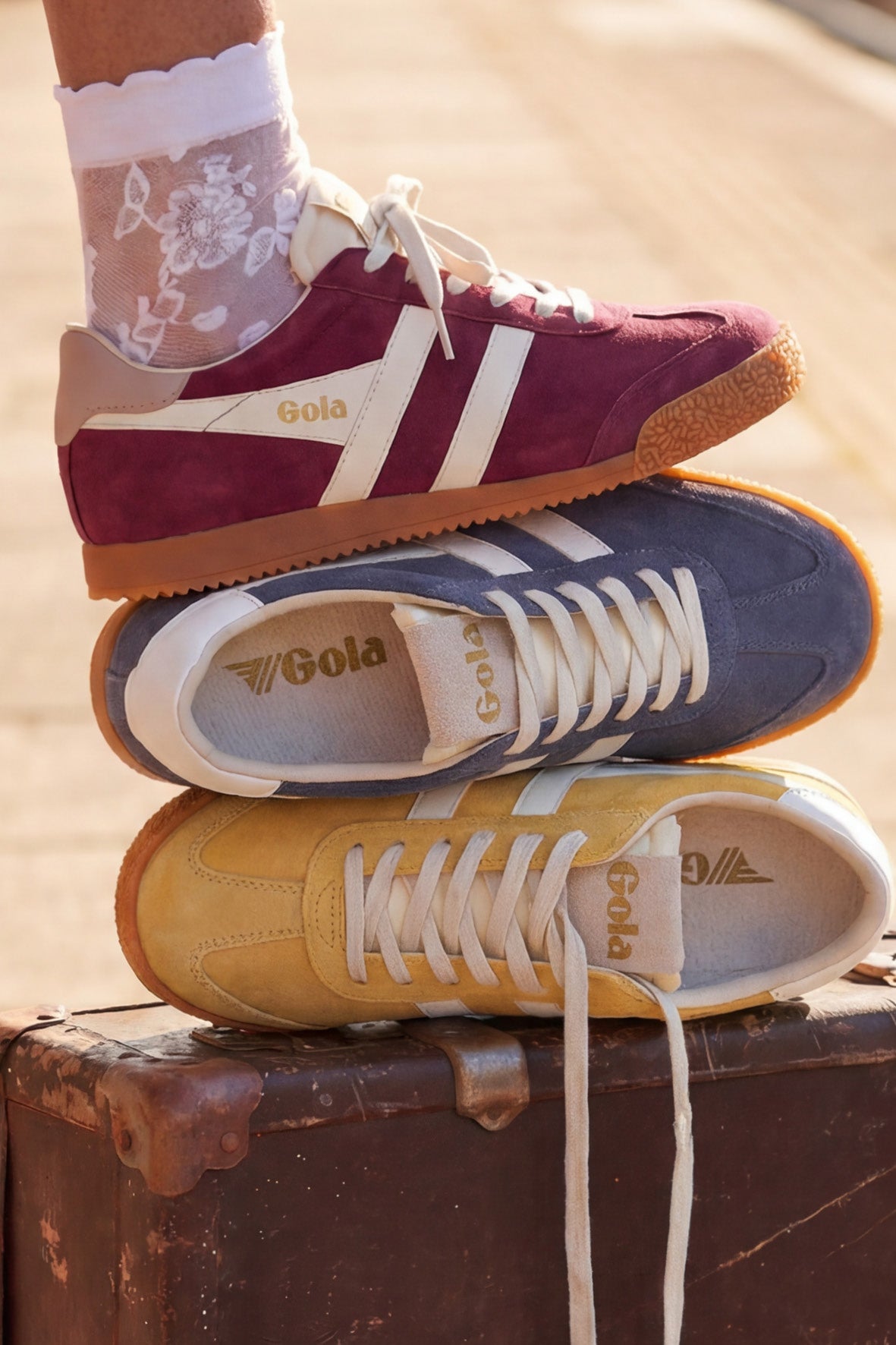 Three Gola trainers stacked on a vintage suitcase. Top shoe is burgundy, middle is navy, bottom is yellow