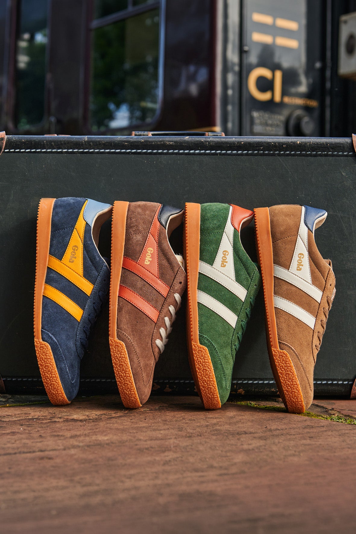 Four Gola Harrier trainers in blue, brown, green, and tan, lined up on a suitcase