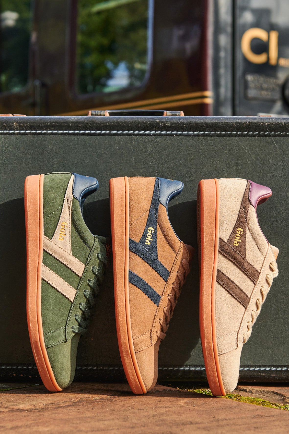 Three Gola trainers in green, tan, and beige, with contrasting stripes and orange soles, on a suitcase