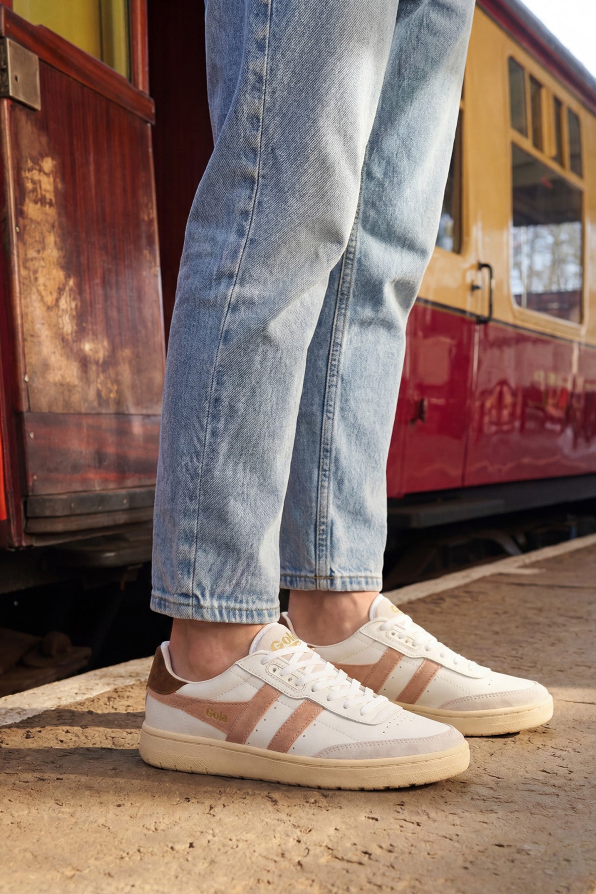 Person wearing light blue jeans and white Gola trainers with pink and brown accents, standing by a train