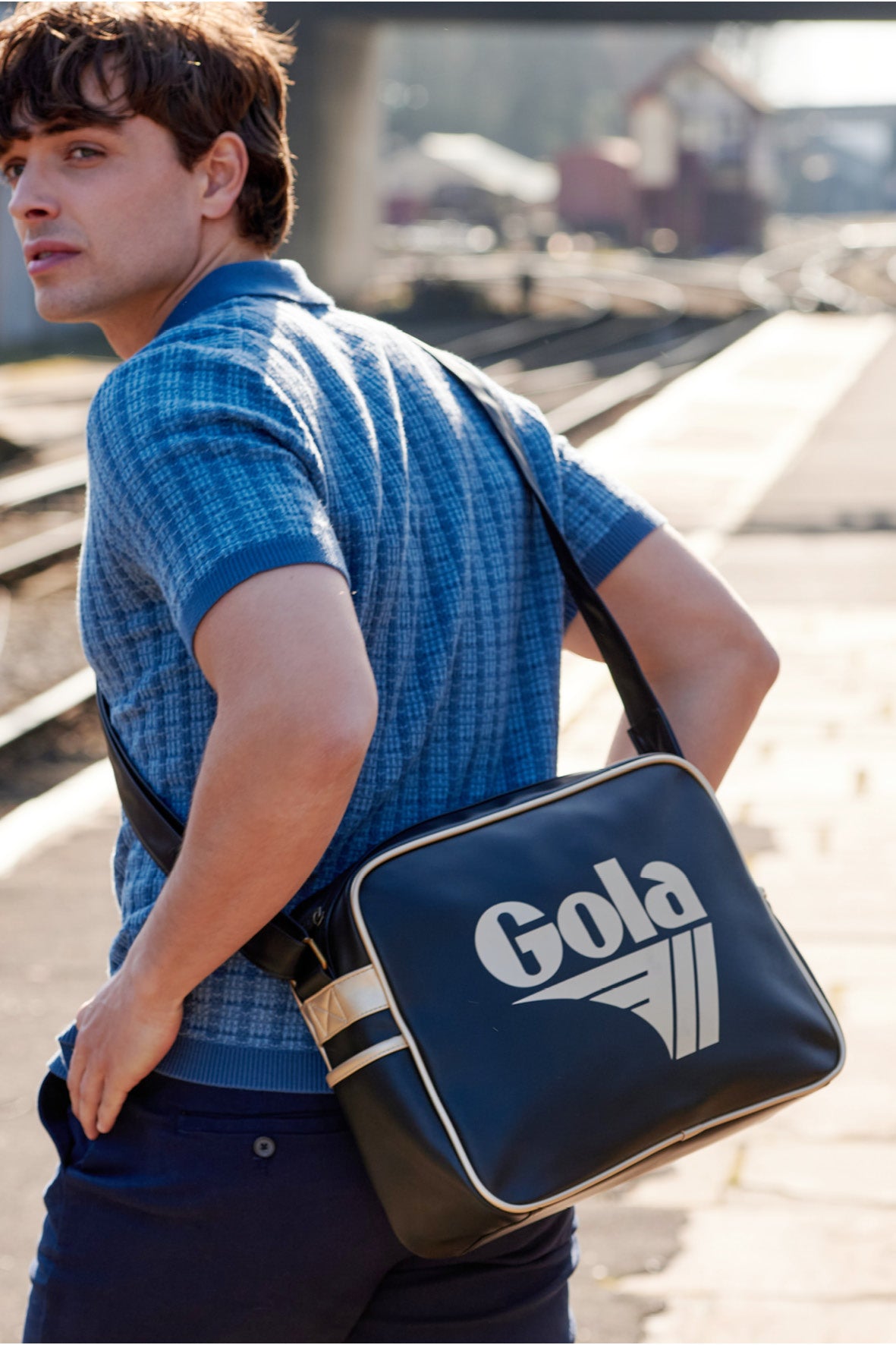 Man in blue polo shirt and navy trousers with a black Gola shoulder bag at a train station