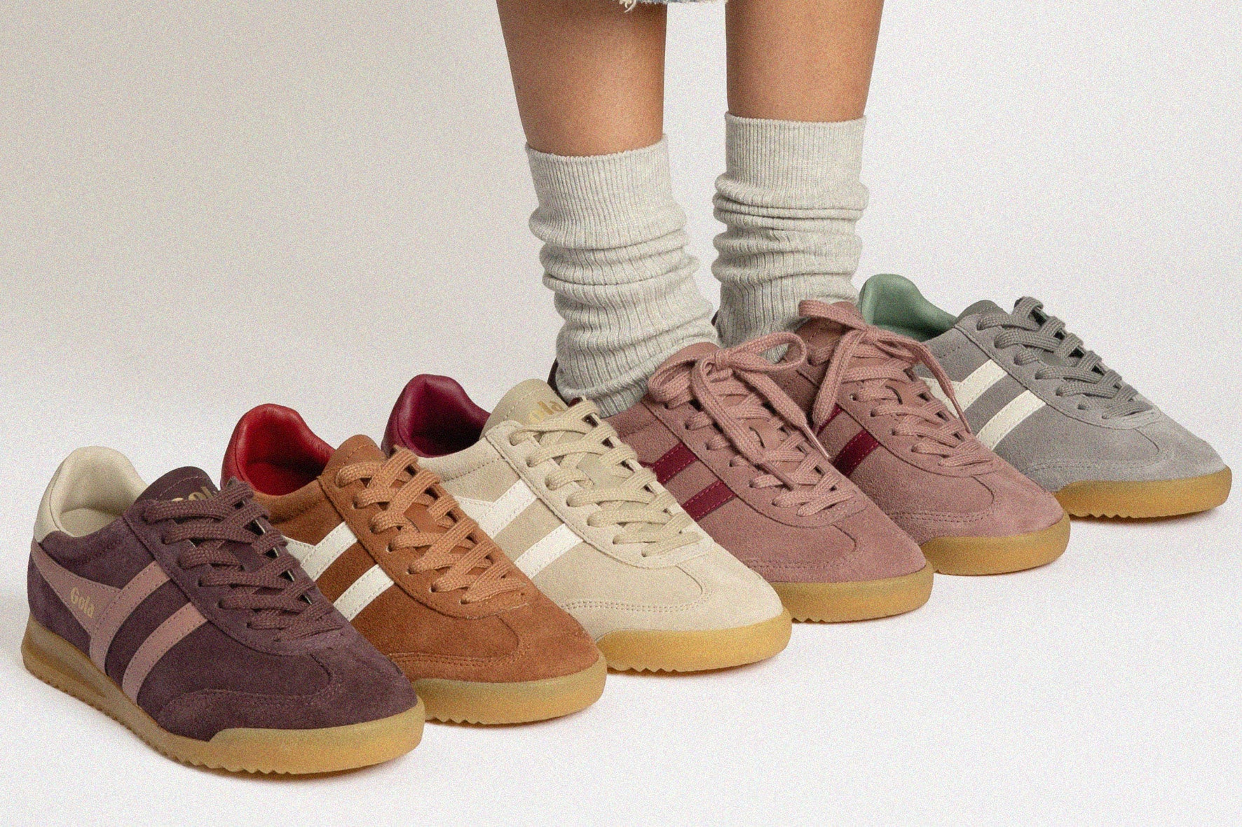 Six suede sneakers in muted earthy tones are lined up on a light gray studio background, with a person's lower legs and socks visible behind them. The shoes have a consistent design with three side stripes, lace-up fronts, and gum soles.