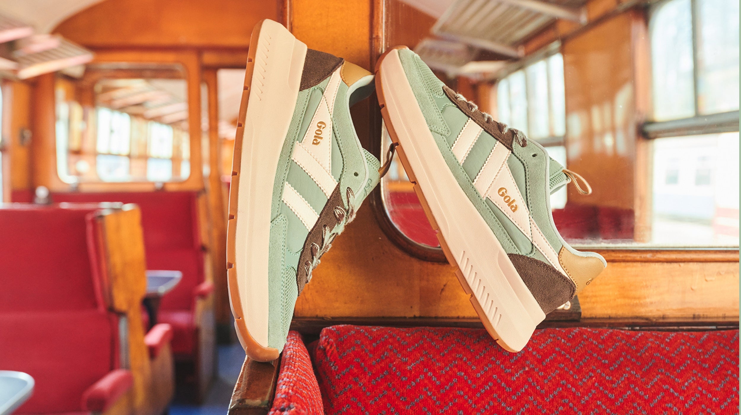 Two Gola sneakers, light green with brown and white accents, rest on a red patterned seat inside a train.