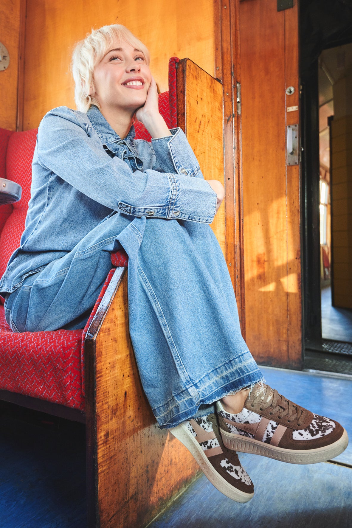Woman in denim outfit and brown and white trainers sitting on a red train seat