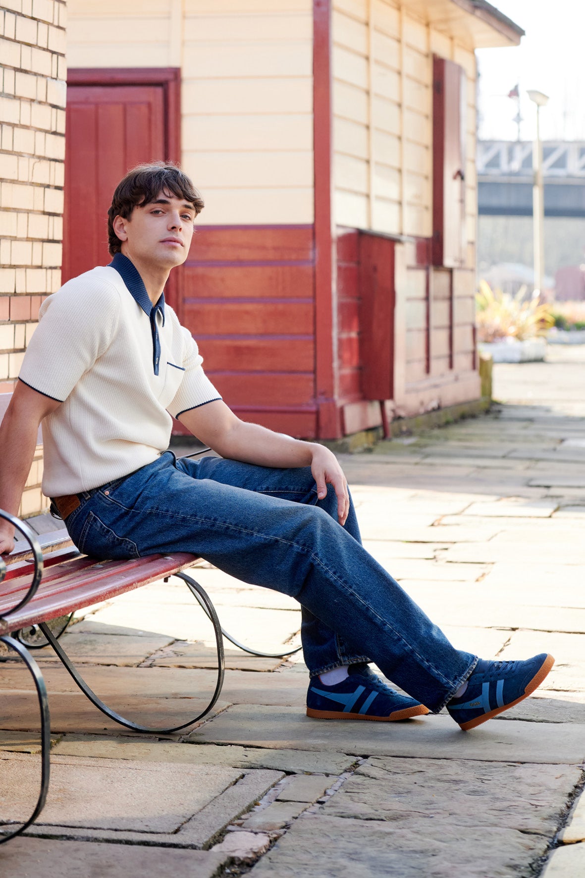 Man in a cream polo shirt, blue jeans, and blue trainers sitting on a red bench