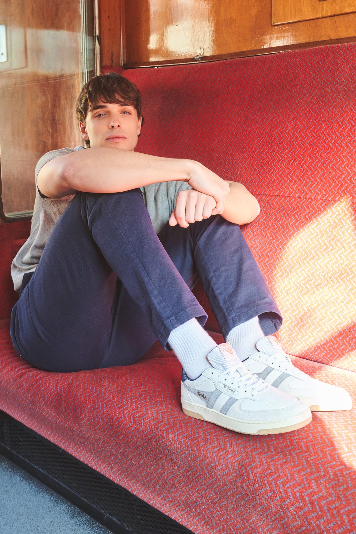 Man in grey t-shirt and navy trousers sitting on a red patterned seat, wearing white Gola trainers