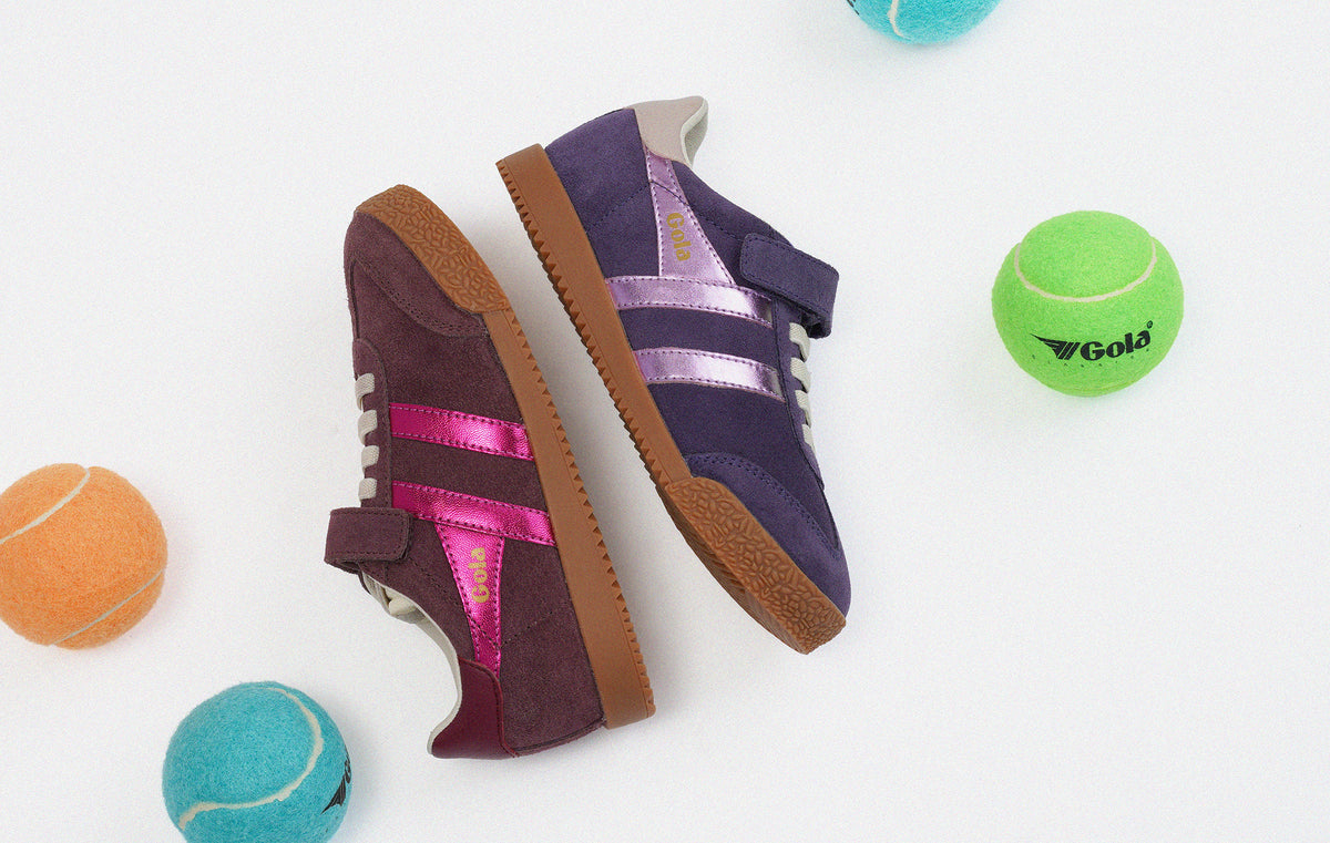 A pair of retro-style low-top sneakers in purple suede with metallic pink and lavender stripes, displayed on a white background with tennis balls around them.
