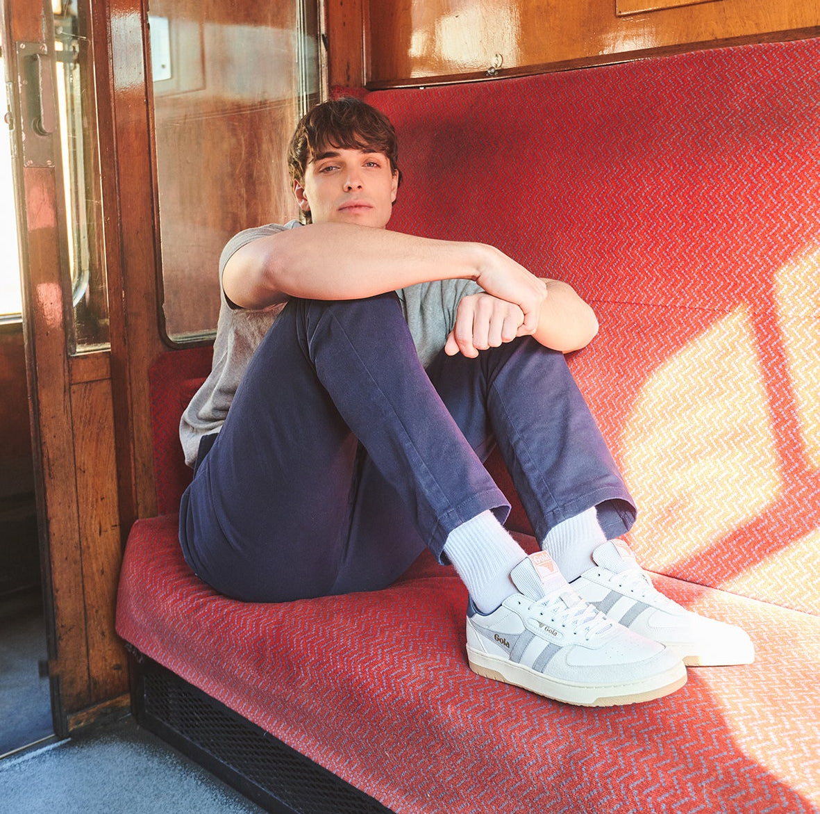 Man in gray shirt and navy pants sitting on a red train seat, wearing white sneakers.
