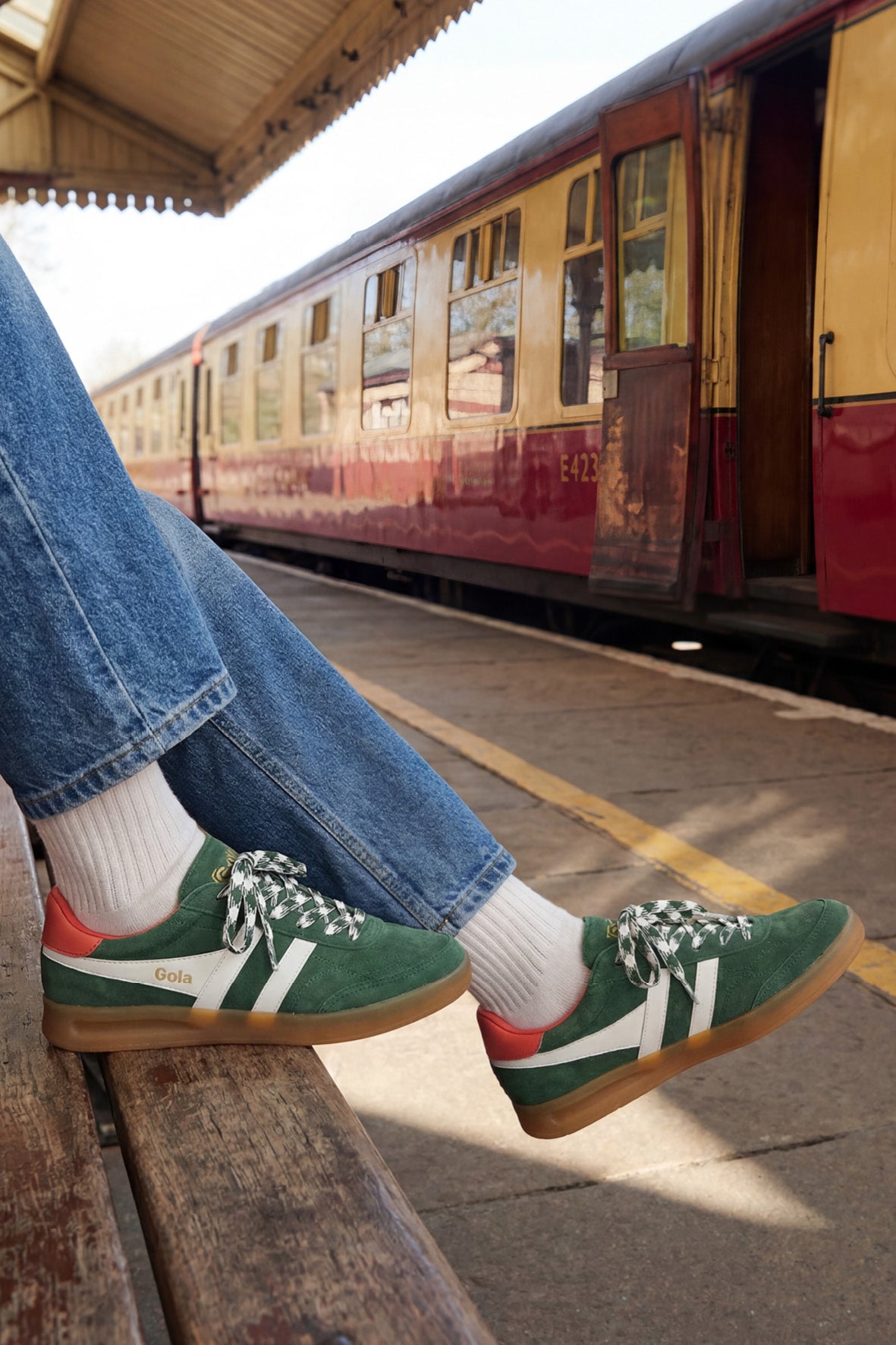 Person wearing green Gola trainers with white stripes and orange heel detail, sitting on a bench at a train station