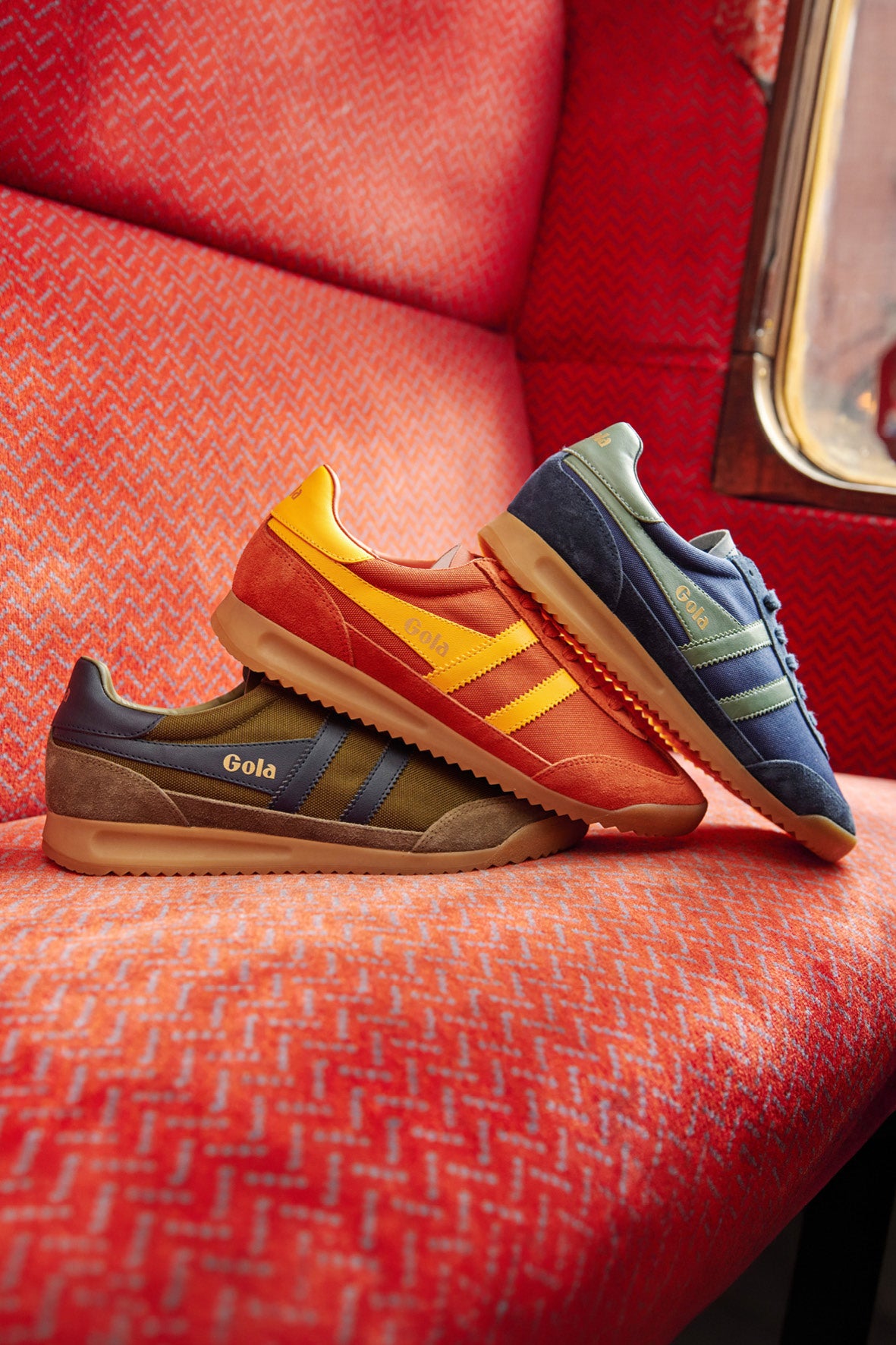 Three Gola trainers in brown, orange, and blue on a red patterned seat