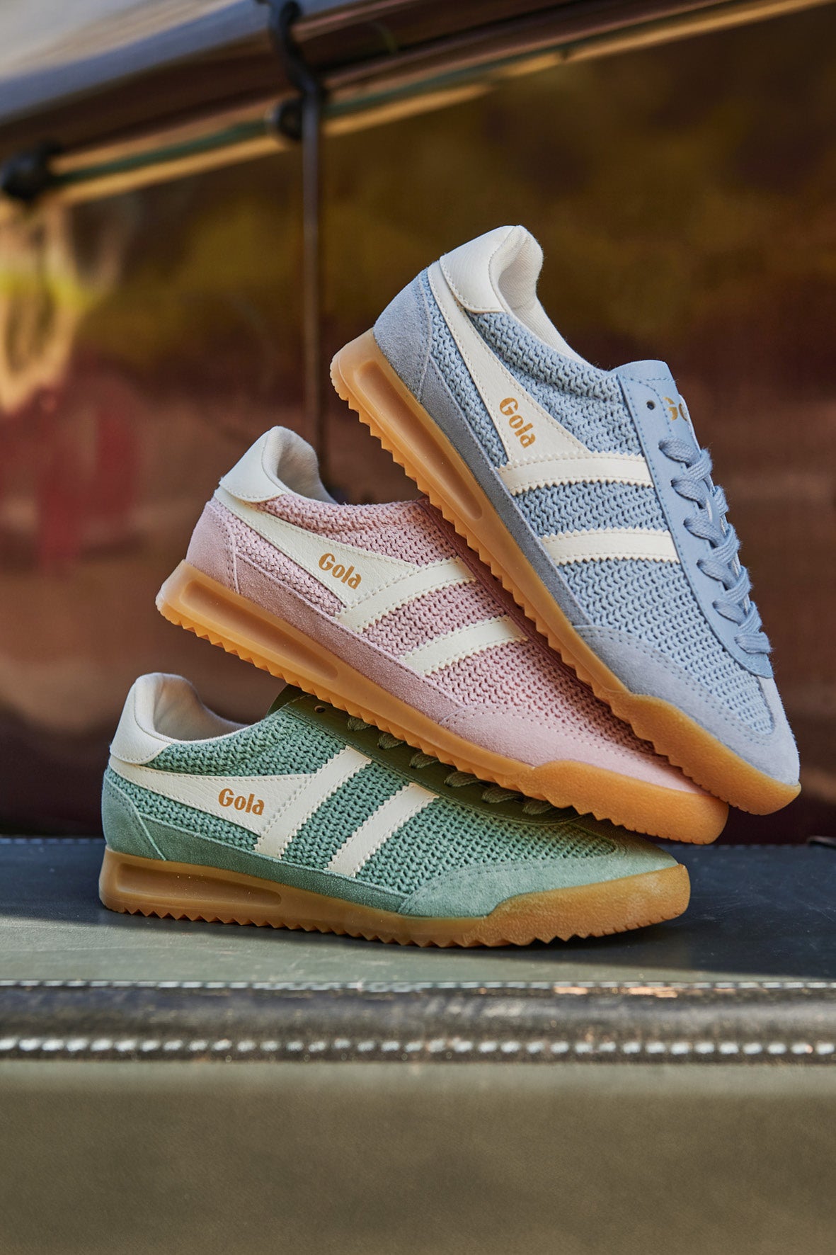 Three Gola trainers stacked, one blue, one pink, one green, all with white stripes and gum soles