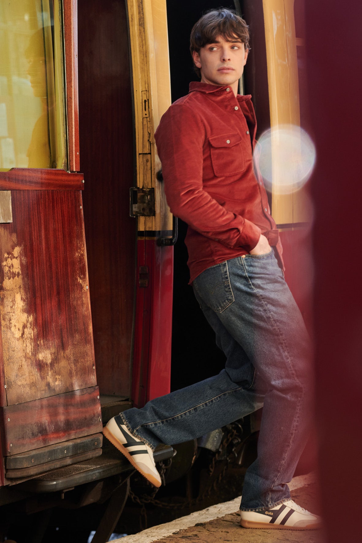 Man in a red corduroy shirt, blue jeans, and Gola trainers stepping off a train