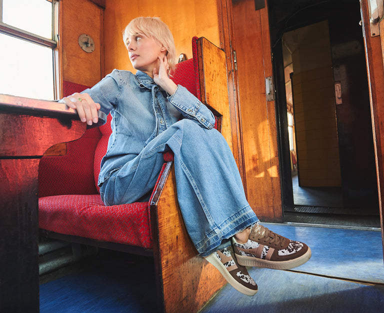 Blonde woman in denim jumpsuit and brown animal print sneakers sitting on a red train seat.