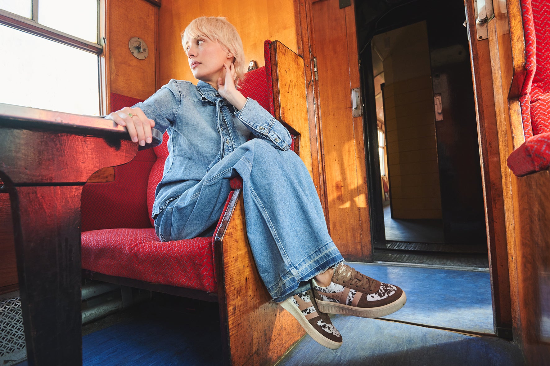 Woman in denim jumpsuit and floral sneakers sits on a red train seat, looking out the window.